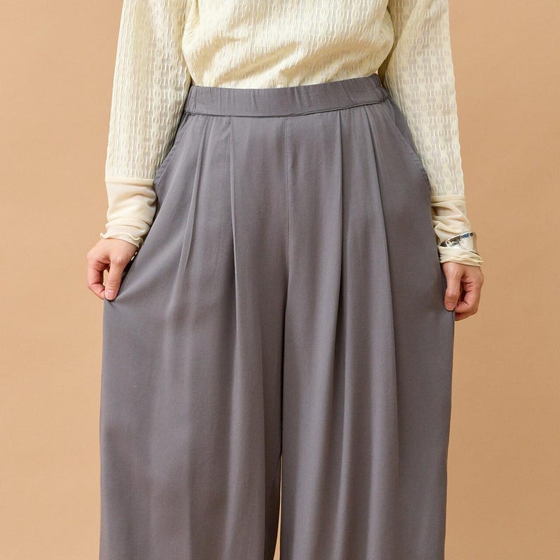 Puede incluir: Pantalones grises de pernera ancha con pliegues en la cintura. Los pantalones est&aacute;n hechos de una tela suave y fluida. La persona lleva una camiseta crema de manga larga.