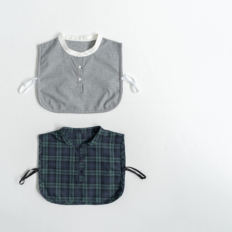 May include: Two fabric collars, one with a white and gray gingham pattern and the other with a green and black plaid pattern. Both collars have a button closure and ties at the sides.