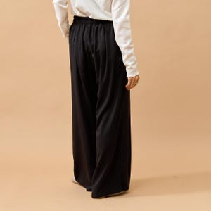 Puede incluir: Pantalones negros de pernera ancha con cintura el&aacute;stica. Los pantalones se combinan con una camisa blanca de manga larga. El conjunto se muestra desde atr&aacute;s, con la persona que lo lleva de pie sobre una superficie de color neutro.
