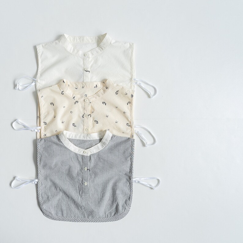 May include: Three sleeveless shirts with different patterns. The top shirt is white, the middle shirt is cream with a small floral pattern, and the bottom shirt is striped in black and white.
