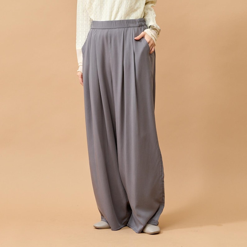 Puede incluir: Pantalones grises de pernera ancha con cintura alta y pliegues. Los pantalones tienen bolsillos y un ajuste holgado y relajado. La modelo lleva una camisa de manga larga color crema. Los pantalones son de tejido suave y fluido.