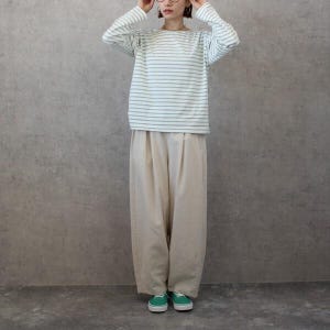 May include: A person wearing a long-sleeved, white and light blue striped shirt, paired with beige, wide-leg pants and green sneakers. The outfit is casual and comfortable, suitable for everyday wear. The background is a plain, gray wall.
