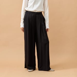 Puede incluir: Pantalones negros de pernera ancha y cintura alta. Los pantalones est&aacute;n hechos de una tela suave y fluida. La modelo lleva una camisa blanca de manga larga. El fondo es de color beige liso.