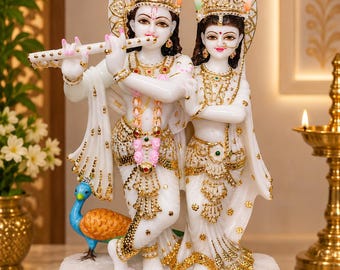 May include: A white marble statue of two figures standing on a rectangular base. One figure plays a white flute, wearing a crown with a green peacock feather. The other figure stands beside them. Both are adorned with gold-painted jewelry and clothing.