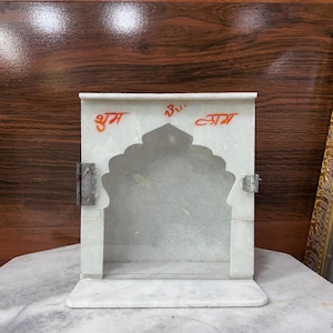 May include: A white marble temple with an arched opening and a rectangular base. The temple has a hinged door and is adorned with red text. The background is a dark wood surface.