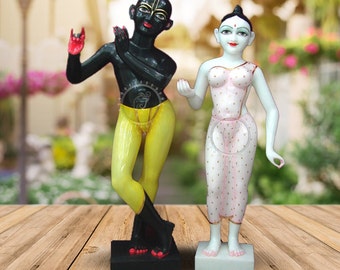 May include: A pair of hand-painted statues depicting Radha and Krishna. Radha is dressed in a white and pink outfit, while Krishna is wearing a yellow dhoti and a black upper body. The statues are standing on separate black and white bases.