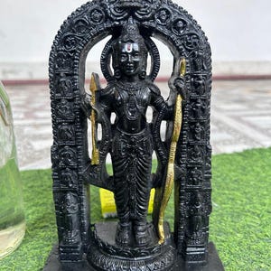 May include: A black deity statue, set within a detailed arch. The figure holds a bow and arrow, with gold detailing. The statue rests on a rectangular base, against a green surface.