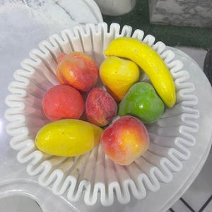 May include: A white, wavy-edged bowl filled with colorful, artificial fruit. The arrangement includes a yellow banana, lemons, peaches, and a green pear. The bowl sits on a white marble surface, creating a decorative display.