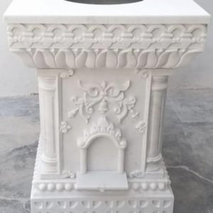 Puede incluir: Un pedestal de mármol blanco con intrincadas tallas. La parte superior cuadrada tiene una abertura circular. Los lados presentan diseños de relieve floral y arquitectónico, incluyendo columnas y un nicho arqueado. La base tiene un patrón geométrico.