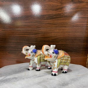 May include: Two white elephant figurines with ornate, colorful detailing. Each elephant has a raised trunk, blue beaded head covering, and decorative patterns in gold, red, and green. The elephants stand on a marble surface.