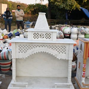 May include: A white marble temple structure with intricate carvings. The design includes a peaked roof, arched details, and a lower platform. The temple is surrounded by various ceramic pots and other decorative items.