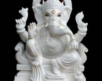 May include: A white marble statue of the deity Ganesha seated on an ornate throne. The figure has four arms, an elephant head with gold accents on the forehead and eyes, and is positioned on a rectangular base with a small mouse figure at the side.
