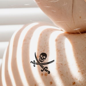 Może przedstawiać: Zbliżenie na ramię osoby z czarno-białym tatuażem czaszki i skrzyżowanych kości.