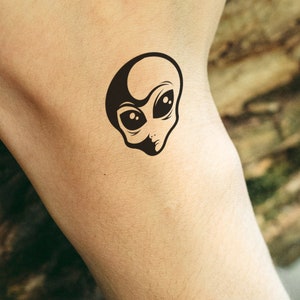 May include: A black alien head temporary tattoo. The alien has large oval eyes and a small mouth. The tattoo is applied to the skin. The design is a simple, yet striking, illustration of an extraterrestrial.
