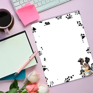 May include: A white sheet of paper with a black and gray paw print border. A black and white border collie puppy is in the bottom right corner holding a birthday cake with lit candles.