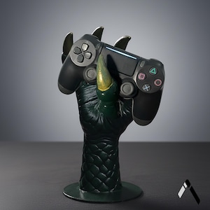 Può includere: Un controller nero per PlayStation 4 tenuto da una statua di mano di mostro verde con artigli dorati. Il controller ha pulsanti colorati e una barra luminosa. La statua è su una base verde scuro.