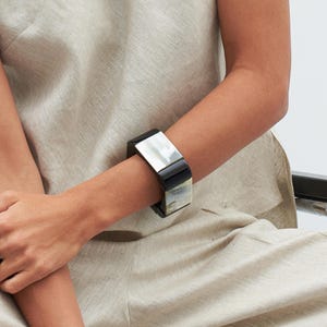 Peut inclure: Un bracelet en corne noir et blanc de forme carrée. Le bracelet est porté au poignet.