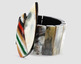 May include: A multi-coloured horn bracelet with a large, striped horn charm. The bracelet is made of multiple pieces of horn, each with a different pattern and colour. The charm is attached to the bracelet with a black elastic band.