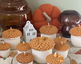 May include: A collection of autumn-themed decorative items, including acorn-shaped candles with brown tops and white bases, a plush orange pumpkin, and a brown glass mushroom. The candles are arranged on a wooden slice, with a fall-themed tablecloth in the background.