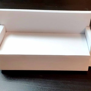 May include: A miniature white sofa with a rectangular back and armrests. The small couch is made of white cardboard and is designed to be a dollhouse furniture item. The sofa is placed on a dark surface.