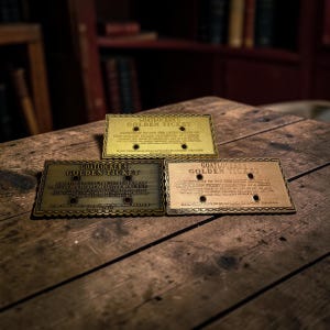 Könnte beinhalten: Drei rechteckige Metallschilder mit gewelltem Rand, jeweils graviert mit dem Text GOATLOCKER'S GOLDEN TICKET. Die Schilder verfügen über vier kreisförmige Löcher und detaillierte Inschriften in verschiedenen Metalloberflächen aus Gold, Bronze und Messing.