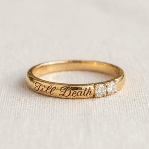 May include: A gold band ring featuring a hammered texture and the engraved script text Till Death. The ring is accented with two small, round, clear, faceted stones set side by side on the band, creating a subtle and elegant appearance.
