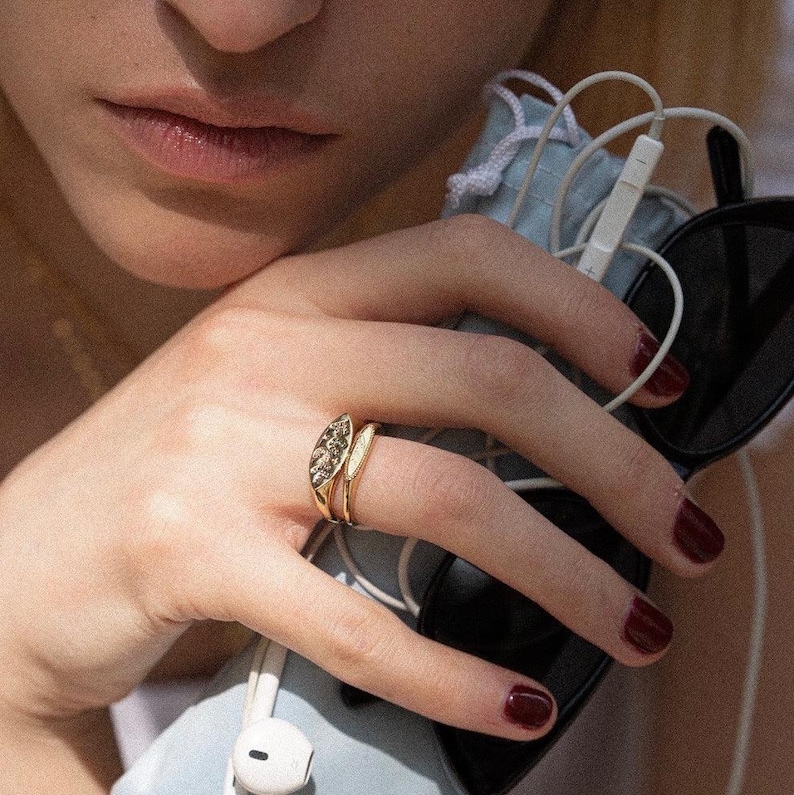 May include: A gold ring featuring an elongated, marquise-shaped face with an engraved celestial star pattern, worn alongside a simple, thin gold band. The ring is displayed on a finger with dark red nail polish, near black sunglasses and white earbuds.