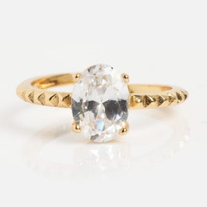 May include: A gold-toned ring featuring a large, clear, oval-cut faceted stone held by four prongs. The band has a textured surface with a repeating pattern of small, triangular indentations along the outer edges on both sides of the center stone.