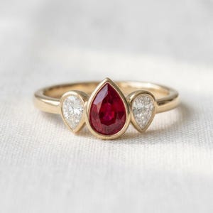 Pode incluir: Um anel de ouro com um rubi vermelho central em lapidação pera, ladeado por dois diamantes brancos menores também em lapidação pera. As pedras estão cravadas numa banda de ouro polido, criando um design de três pedras com rubi vermelho.