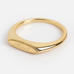 May include: A gold-toned ring featuring a smooth, elongated oval face with a beaded border. The center of the oval face is engraved with the word Bitok in a cursive script. The band is polished and tapers toward the back of the ring.