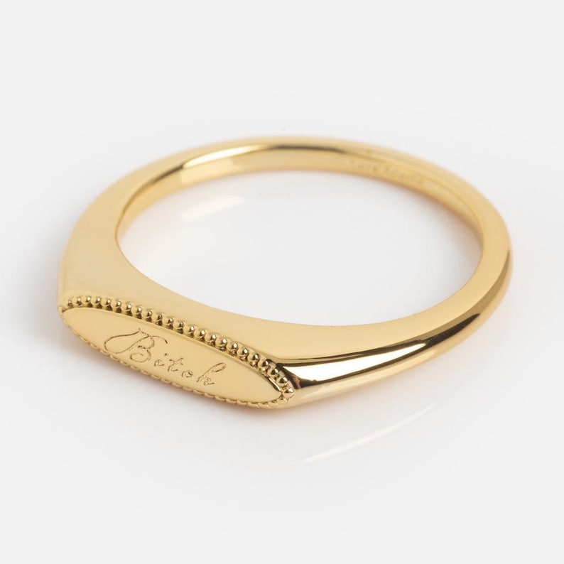 May include: A gold-toned ring featuring a smooth, elongated oval face with a beaded border. The center of the oval face is engraved with the word Bitok in a cursive script. The band is polished and tapers toward the back of the ring.