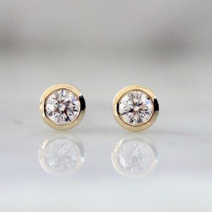 May include: A pair of round stud earrings featuring clear, faceted stones set in polished yellow gold bezels. The earrings are circular in shape with a smooth, metallic border surrounding each brilliant-cut stone, reflecting light clearly.