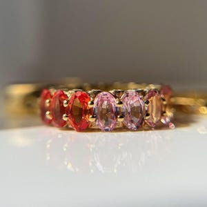 May include: A gold ring featuring a row of oval-cut gemstones in shades of red, pink, and peach. The gemstones are set closely together, creating a continuous band of color. The ring is displayed on a white surface.