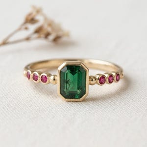 May include: A gold ring featuring a central emerald-cut green gemstone in a bezel setting, flanked by three small round-cut red gemstones on each side, set within a band characterised by a series of small gold spheres along the shank.