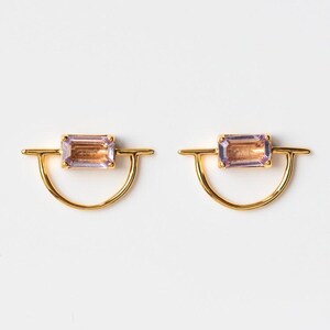 May include: A pair of gold-colored earrings. Each earring features a rectangular, light purple gemstone set in a gold frame. The frame has a curved, half-circle design with a horizontal bar extending from each side of the gemstone.