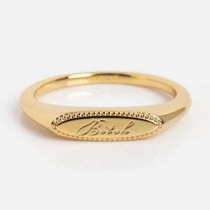 May include: A gold-toned ring featuring an oval-shaped face with a beaded border. The center of the oval is engraved with the word "Bitch" in a cursive script font. The band is smooth and tapers slightly toward the back of the ring.