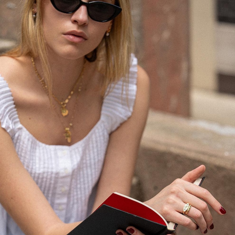 May include: A white sleeveless top with a textured grid pattern, layered gold necklaces, black cat-eye sunglasses, and a gold ring with a central stone worn on the finger while holding an open book with a black cover and red interior pages.