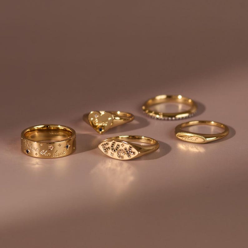 May include: A set of five gold-toned rings. One ring features the text "What the fuck" with small star accents. Another ring displays the word "FUCK" in a central oval setting. A third ring has a heart shape, while two others are plain bands.
