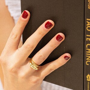 May include: A gold-toned metal ring featuring a flat, rectangular face with the word "FUCK" engraved in black lettering. The ring is worn on a finger with dark red nail polish, positioned next to a dark brown book with gold text on the spine.