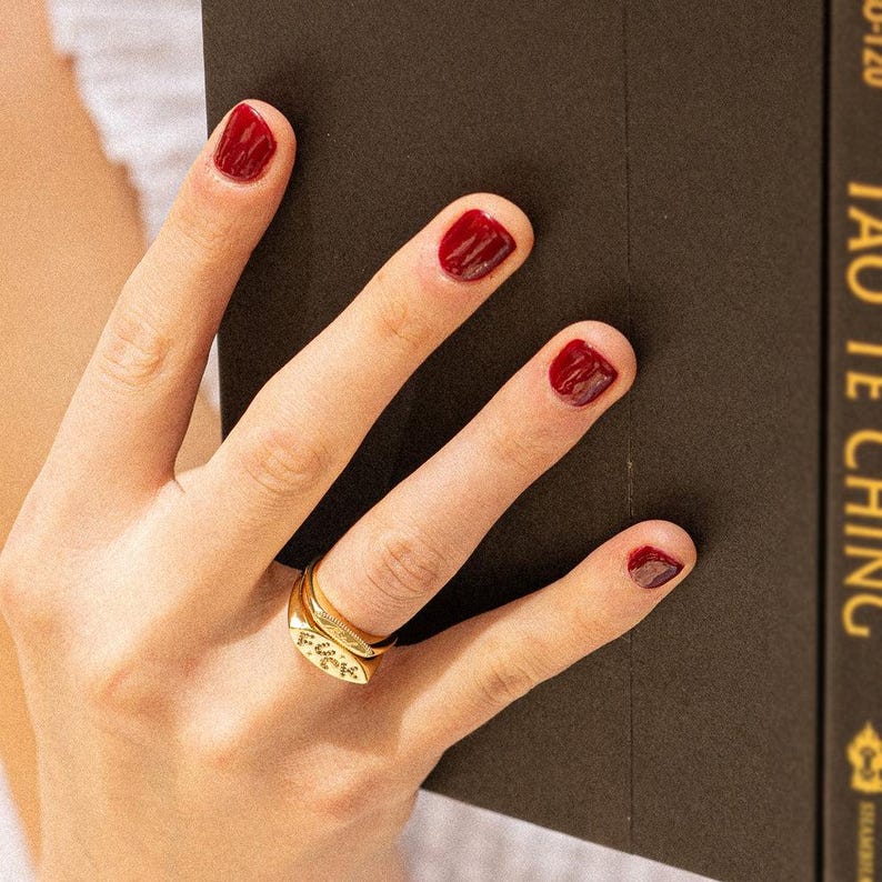May include: A gold-toned metal ring featuring a flat, rectangular face with the word "FUCK" engraved in black lettering. The ring is worn on a finger with dark red nail polish, positioned next to a dark brown book with gold text on the spine.