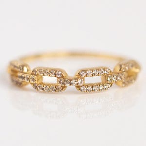 May include: A gold chain link ring with small, sparkling stones set into each link. The ring is displayed on a white surface, reflecting the light and highlighting the ring's intricate design. The ring is a delicate piece of jewelry.