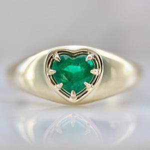 May include: A gold ring featuring a heart-shaped, vibrant green gemstone. The gemstone is set within a heart-shaped frame, and the ring has a smooth, polished band. The ring's design is elegant and eye-catching.