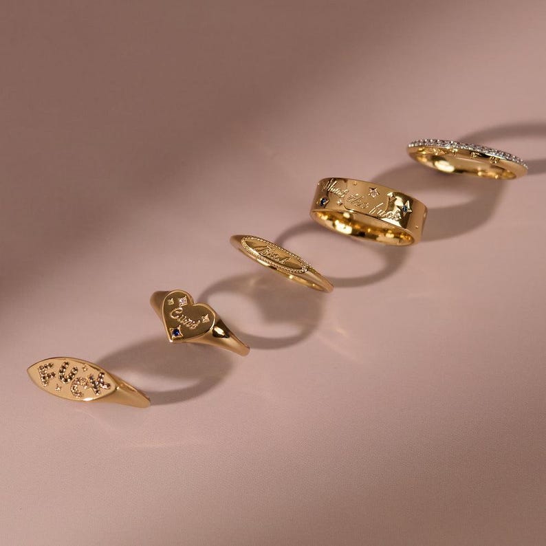 May include: A collection of five gold-toned rings arranged in a diagonal line. The rings feature various designs including an oval band with the word FUCK, a heart-shaped ring with the word Cunt, a thin band, a wide band with text, and a pav&eacute; band.