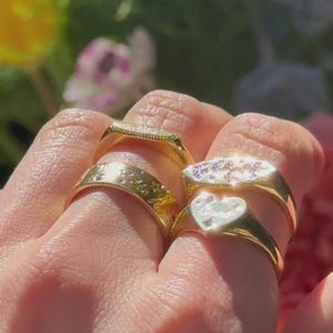 May include: A collection of four gold-toned rings worn on fingers. One ring features a heart-shaped face, another has an oval face with small embedded stones, and two are simple bands with engraved star-like patterns on the outer surface.