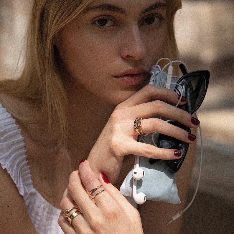 May include: A set of gold-toned rings worn on fingers, featuring various band designs including a textured piece and a slim band with small embedded stones. The rings are displayed on fingers that are also holding black sunglasses and white wired earbuds.