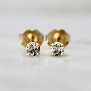 May include: A pair of stud earrings featuring round, salt and pepper diamonds set in four-prong yellow gold settings. The earrings have a classic post-back design, showcasing the speckled grey and black inclusions within the circular stones.