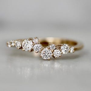 Könnte beinhalten: Ein goldfarbener Ring mit einer Ansammlung runder, klarer Edelsteine. Die Edelsteine sind unterschiedlich groß und eng beieinander gesetzt, wodurch ein funkelndes, asymmetrisches Design entsteht. Der Ring ist glatt und poliert.