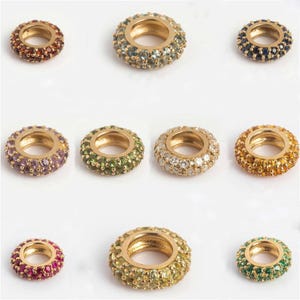 May include: A collection of gold-toned, circular beads, each encrusted with a variety of colorful gemstones. The beads feature a central hole and are arranged on a white background. Gemstone colors include red, green, blue, purple, and yellow.