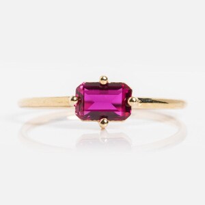May include: A gold ring featuring a rectangular, vibrant pink gemstone. The gemstone is set with four small gold accents. The ring's band is a thin, polished gold. The ring is displayed against a white background.