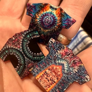 May include: Three miniature tie-dye t-shirts in shades of blue, purple, orange, and green are held in a hand. Each shirt features a unique swirling pattern. The shirts are small, likely intended as charms or decorative items.